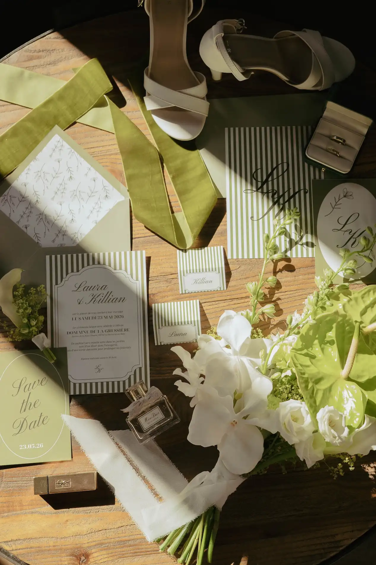 Mise en scène d'accessoires de mariage sur une table en bois, incluant le faire-part de Laura et Killian, des alliances, des chaussures blanches, un parfum Miss Dior et un bouquet de fleurs blanches et vertes.