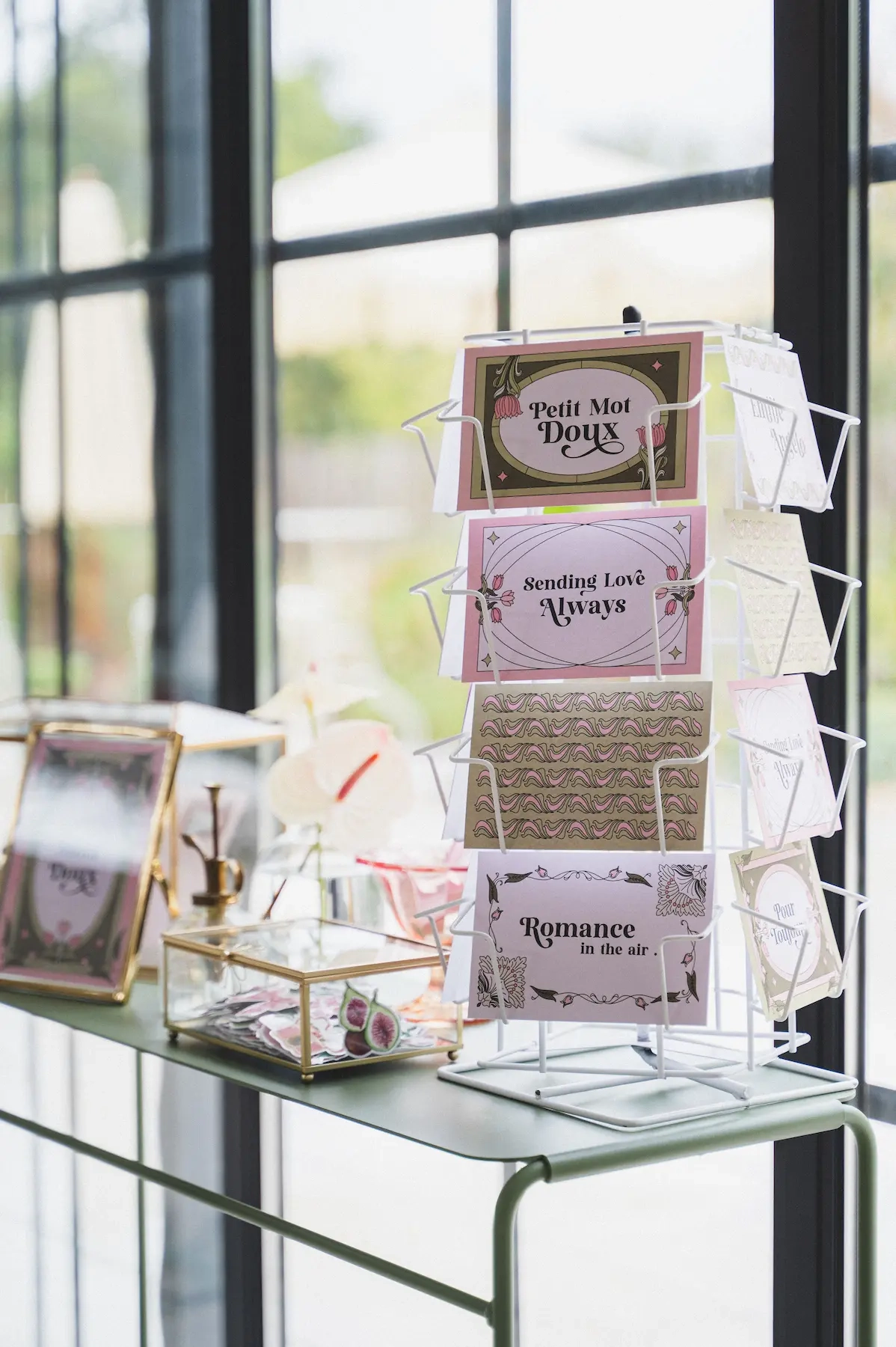 Un présentoir rotatif en métal blanc expose des cartes postales décorées sur le thème du mariage, avec des inscriptions comme « Petit Mot Doux », « Sending Love Always » et « Romance in the air ». L'ensemble est disposé sur une table près d'une fenêtre, aux côtés d'une boîte en verre contenant d'autres cartes. 