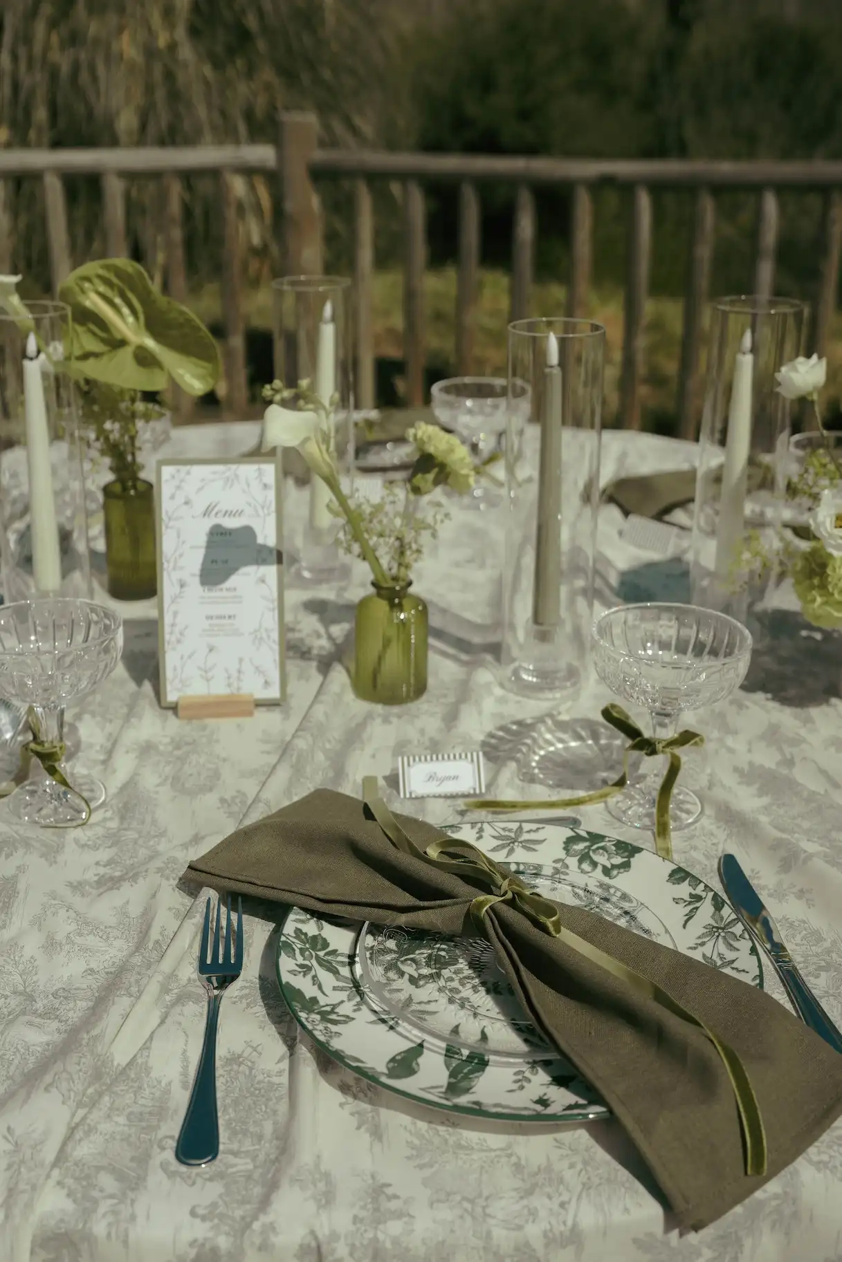 Table de mariage dressée avec une nappe à motifs, des assiettes décorées de motifs végétaux, une serviette en tissu vert olive nouée d'un ruban et un menu imprimé. La décoration comprend des bougies dans des photophores en verre et de petites fleurs dans des vases.