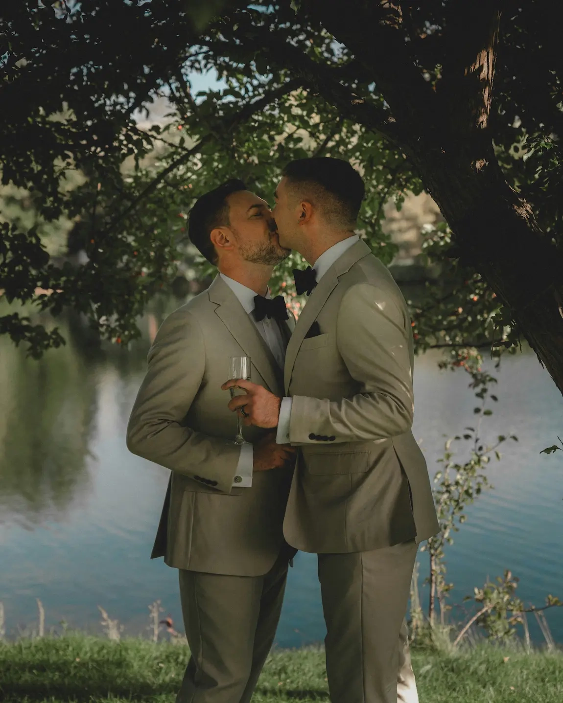 Deux hommes en costume vert clair et nœud papillon noir s'embrassent tendrement sous un arbre au bord d'un lac, l'un d'eux tenant une flûte de champagne. 