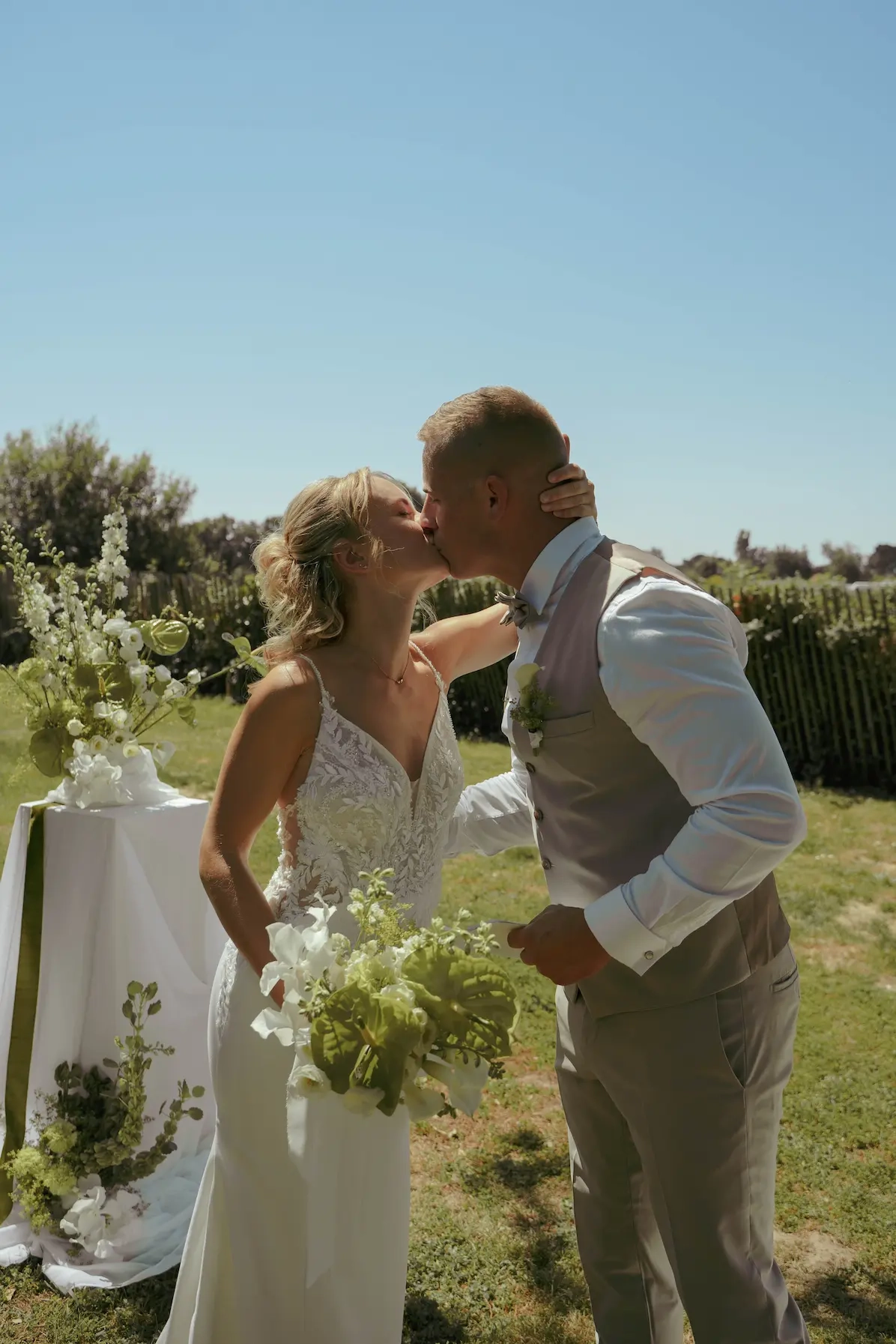 Une mariée en robe blanche et un marié en costume beige s'embrassent tendrement lors d'une cérémonie en extérieur. La mariée tient un bouquet de fleurs blanches et vertes dans un cadre champêtre ensoleillé.