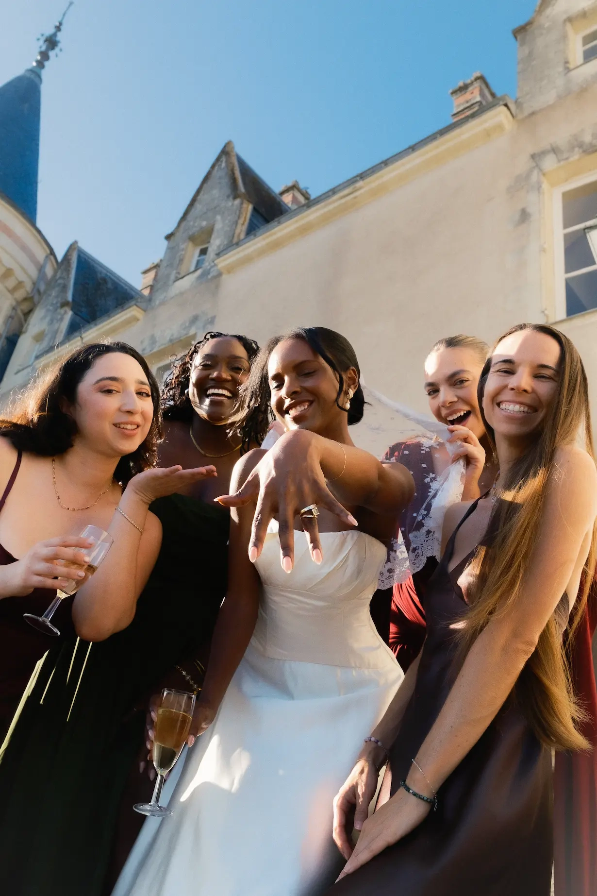 La mariée, entourée de ses amies lors du cocktail, présente fièrement son alliance à l'objectif. Toutes sourient devant un château sous un ciel ensoleillé. 