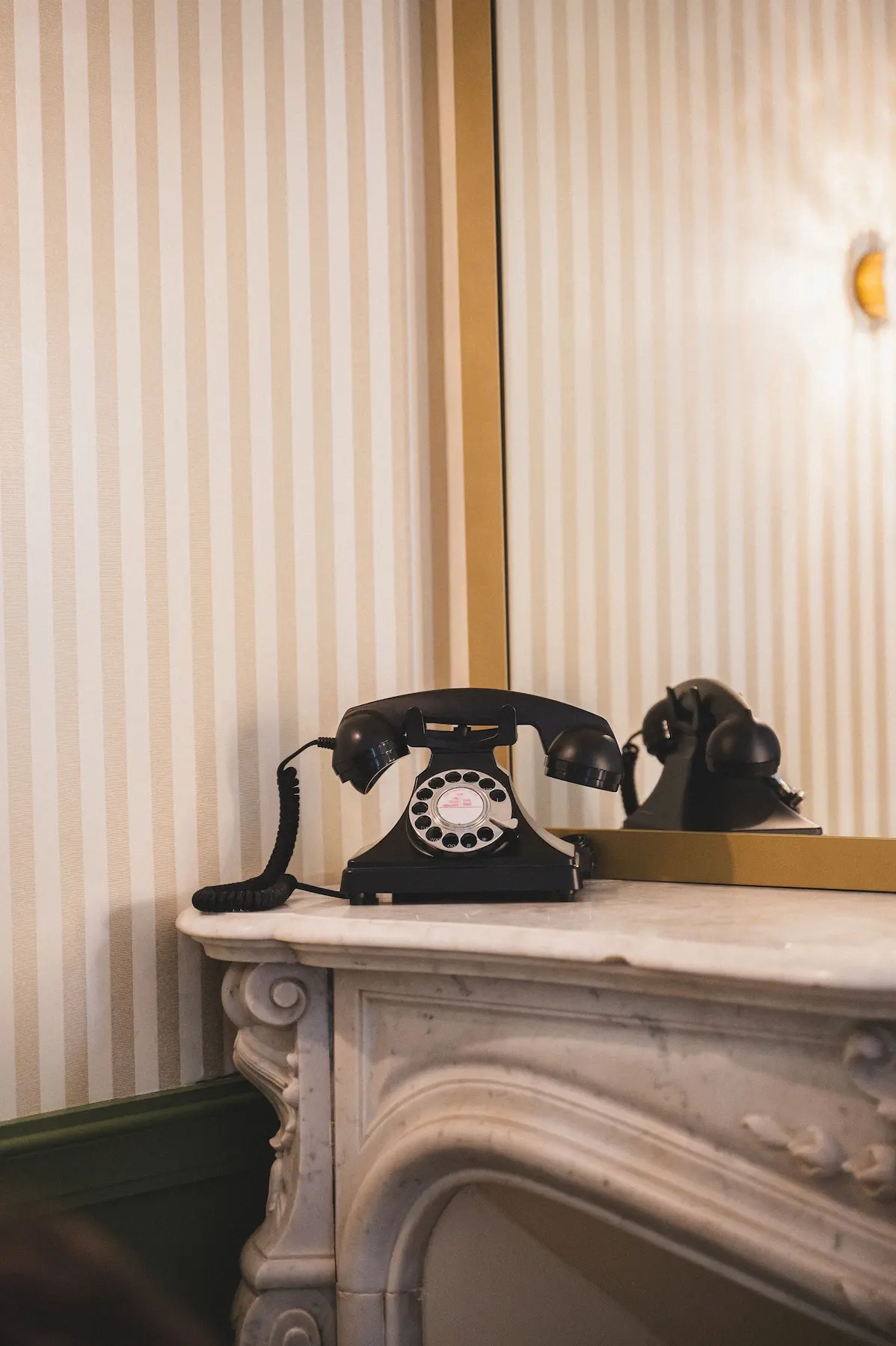 Un téléphone à cadran noir est posé sur une cheminée en marbre blanc, devant un miroir et un mur au papier peint rayé. 