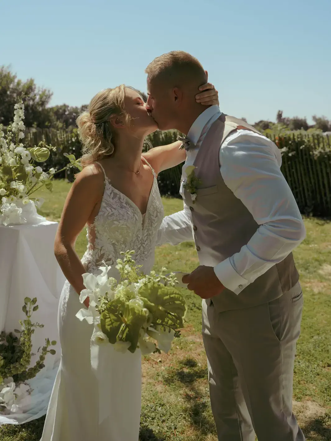 Une mariée en robe blanche et un marié en costume beige s'embrassent tendrement lors d'une cérémonie en extérieur. La mariée tient un bouquet de fleurs blanches et vertes dans un cadre champêtre ensoleillé.