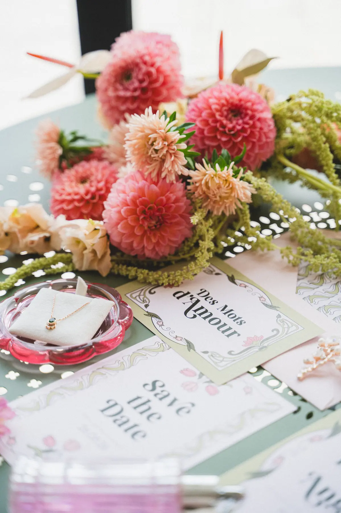 Mise en scène élégante d'accessoires de mariage sur une table en fer verte, incluant de la papeterie dont le Save-the-Date d’Emilie et Angelo, une carte postale, un collier serti d’un pierre verte, une broche doré, un parfum rose et un bouquet de fleurs roses et vertes.
