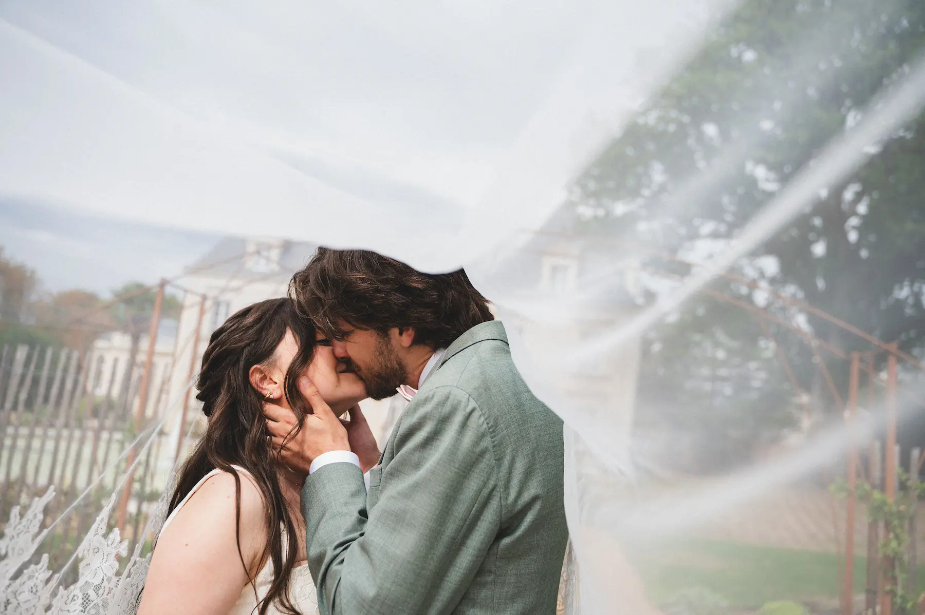 Un couple de jeunes mariés s'embrasse tendrement, partiellement recouvert par le voile de la mariée qui flotte au premier plan. 
