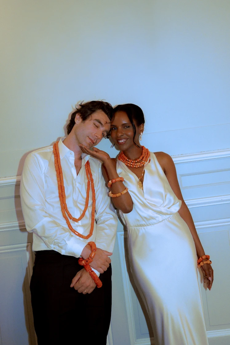 Un couple de jeunes marié·es pose ensemble, parés de colliers et de bracelets en perles de corail orange. La femme, en robe blanche, soutient tendrement le visage de son époux, qui porte une chemise blanche et un pantalon sombre.