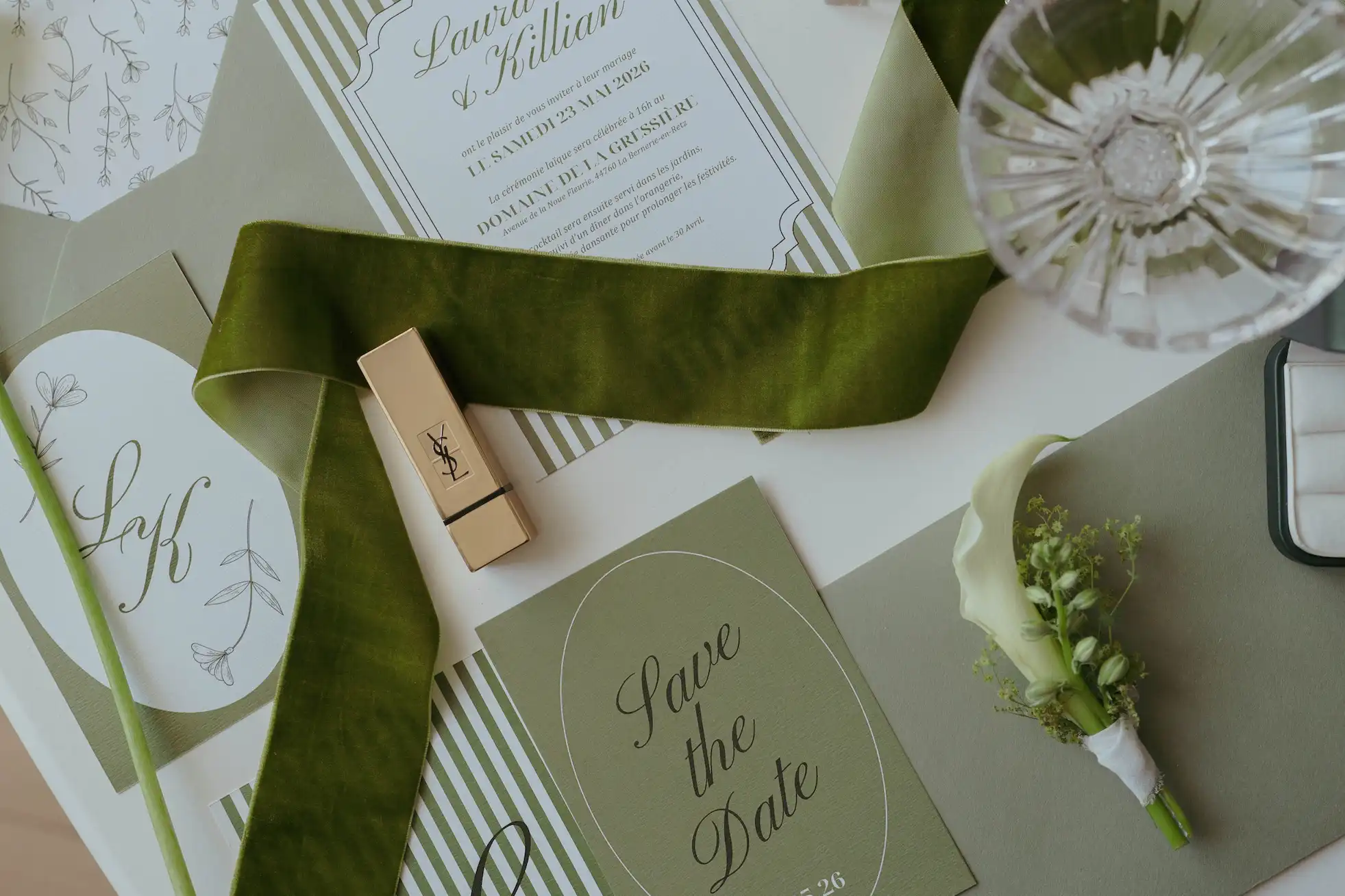 Faire-part de mariage de Laura et Killian pour le samedi 23 mai 2026 au Domaine de la Gressière, accompagné d'un carton Save the Date, d'un ruban de velours vert, d'un rouge à lèvres Yves Saint Laurent et d'une boutonnière florale.