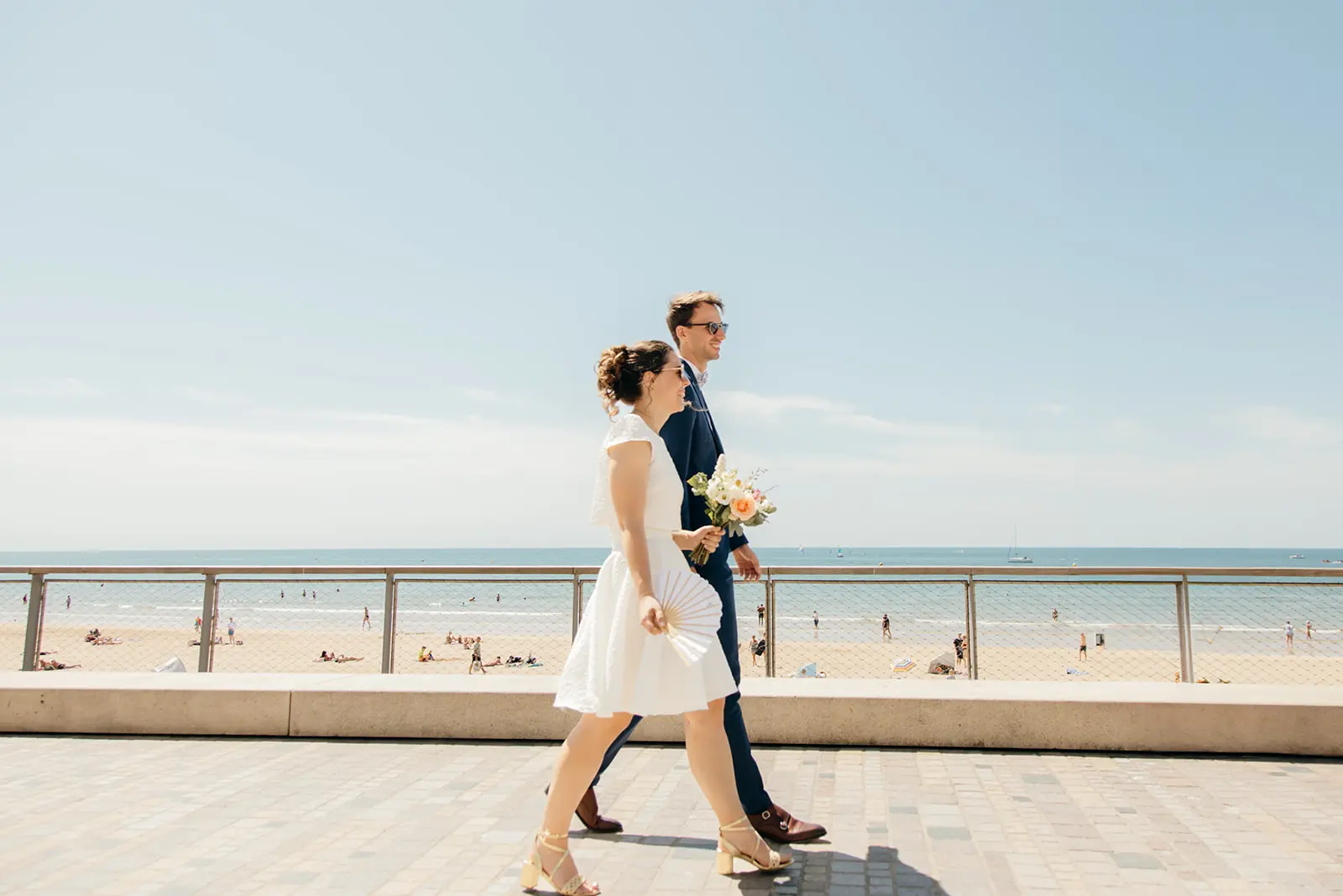Un couple de jeunes mariés marche main dans la main sur une promenade en bord de mer, sous un ciel ensoleillé. La mariée porte une robe blanche courte et tient un bouquet de fleurs, tandis que le marié est vêtu d'un costume bleu marine.
