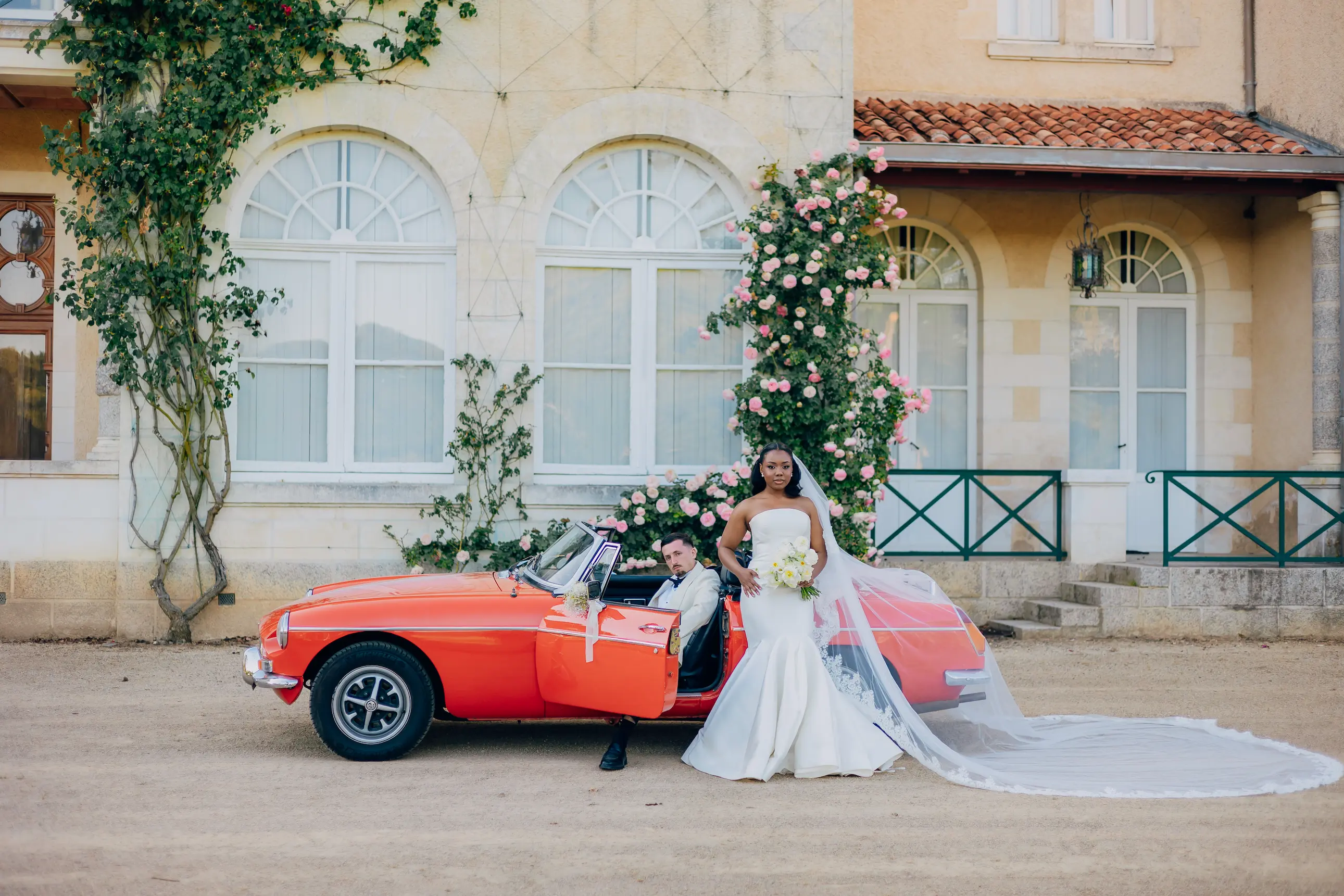 Une mariée en robe blanche avec un long voile et un marié en costume clair posent devant une voiture de collection orange, stationnée devant une demeure ancienne ornée de rosiers.